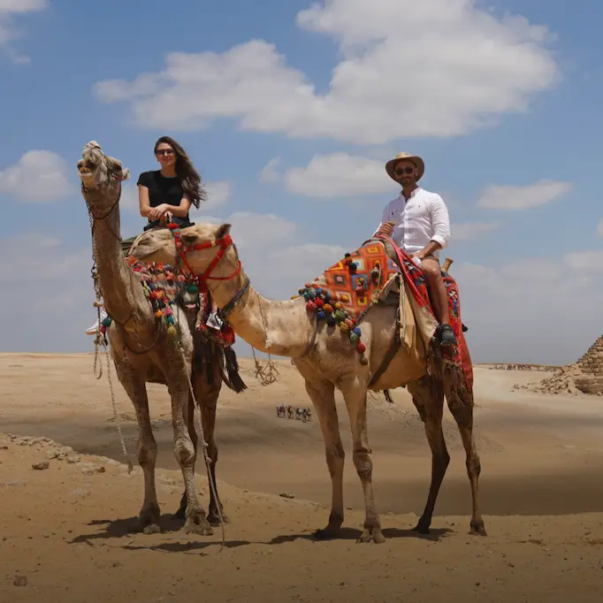 Two people riding camels in the desert, with pyramids in the background
