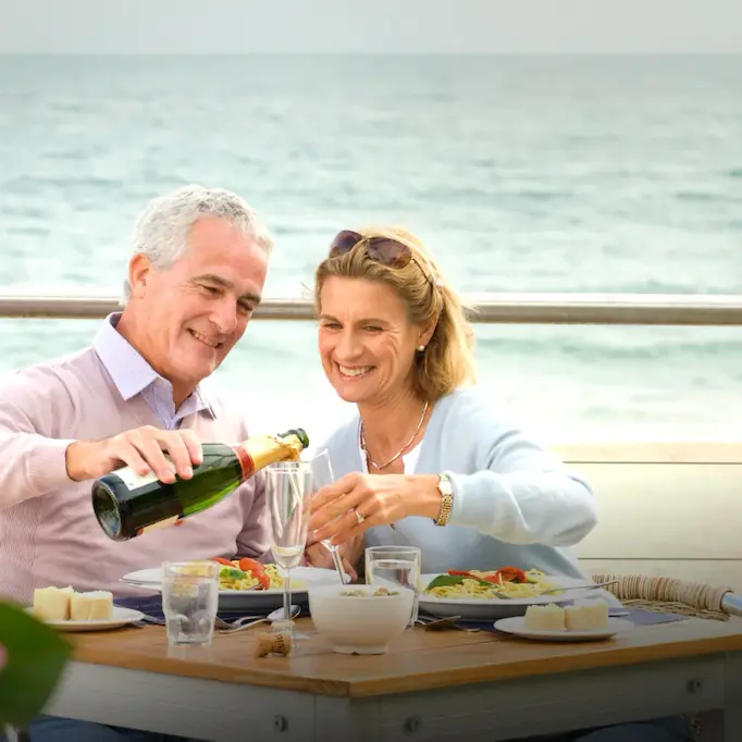 Older couple enjoying a meal and pouring champagne at an outdoor seaside restaurant in France