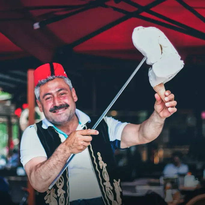 Smiling vendor in traditional attire serving Turkish ice cream.