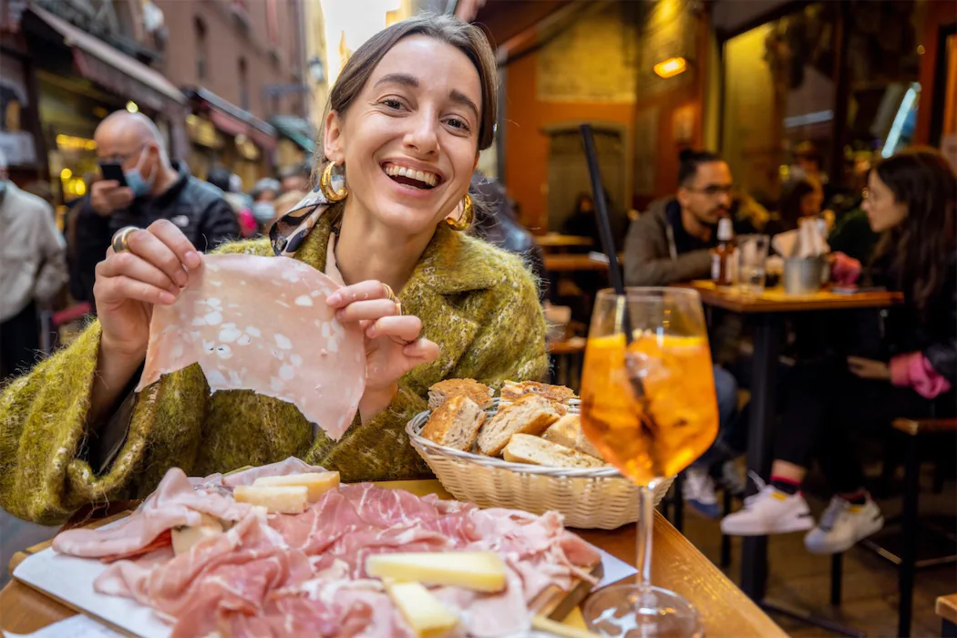 A smiling woman holding a slice of cured ham with a basket of bread and Aperol Spritz at a traditional Catalan charcuterie restaurant in Barcelona.